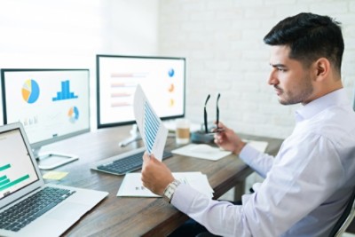 Attractive young male sales professional analyzing chart while holding paper at desk